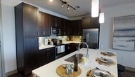 This single-floor residence showcases a thoughtfully designed interior centered around a striking, modern kitchen. The kitchen is a standout feature, boasting rich, dark espresso-toned cabinetry that extends from floor to ceiling, providing generous storage throughout. Crisp white countertops offer a bold contrast against the dark wood tones, creating a visually dynamic and elegant aesthetic. A large kitchen island serves as both a functional workspace and a casual dining area, complete with a deep undermount sink and a sleek, high-arc faucet. Stainless steel appliances — including a French door refrigerator, a smooth-top range, and an over-the-range microwave — lend a professional, contemporary feel to the space. Track lighting overhead and pendant lights above the island ensure the kitchen is well-lit and inviting. Under-cabinet lighting adds warmth and enhances visibility along the work surfaces. The layout is designed for efficiency and flow, with an L-shaped configuration that maximizes counter and cabinet space. The overall ambiance is modern and refined, combining practical design with upscale finishes to create a kitchen that is as functional as it is visually impressive.