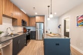 This well-designed single-floor residence offers a thoughtful open-concept layout that seamlessly connects the kitchen, dining, and living areas. The kitchen is a standout feature, outfitted with two-tone cabinetry — warm wood uppers paired with dark navy lowers — complemented by a white subway tile backsplash, stone countertops, and a full suite of stainless steel appliances including a gas range, built-in microwave, dishwasher, and refrigerator. A central island with seating provides an ideal casual dining and gathering space, illuminated by stylish pendant lighting overhead. Light wood-style flooring flows throughout, unifying the open living area, which benefits from large windows that invite abundant natural light and outdoor views. The bedroom is a calm, well-proportioned retreat with large windows, light-toned walls, and wood flooring, creating a bright and restful atmosphere. A spacious walk-in closet with wire shelving offers generous storage. The bathroom features a clean, minimalist design with a soaking tub and shower combo, light tile surround, a dark-cabinetry vanity, and wood-look flooring. The overall aesthetic of the space is modern and cohesive, balancing warm material tones with contemporary finishes.