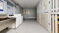 Located at 3017 Queen St E in Toronto's east end, this shared utility space serves as a combined laundry and storage facility. The elongated, single-room layout is finished with durable painted concrete floors and bright white ceilings equipped with fluorescent strip lighting that ensures clear visibility throughout.

Along one side of the room, a row of numbered wooden storage lockers lines the wall, providing residents with individual, slatted-frame storage units. On the opposite side, commercial-grade coin-operated washing machines and dryers are neatly arranged alongside a deep stainless steel utility sink, offering a fully functional laundry setup. A sturdy work table is positioned near the rear of the space, providing a convenient surface for folding or sorting laundry.

Exposed overhead ductwork and piping run along the walls and ceiling, reflecting the practical, utilitarian character of the space. A small window near the laundry appliances allows for some natural light and ventilation. Safety features include a prominently mounted fire extinguisher and clearly marked emergency shutoff signage. A separate electrical room is accessible via a labeled door, and an informational bulletin board is posted on the wall. The overall environment is clean, organized, and purpose-built for everyday residential utility use.
