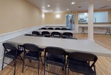 This single-floor multipurpose hall offers a clean, well-appointed space designed to accommodate a variety of gatherings, from meetings and classes to banquets and community events. The open floor plan is anchored by polished light hardwood flooring that extends throughout, lending warmth and a sense of spaciousness to the entire area.

Structural white columns are positioned symmetrically across the room, adding an architectural character while subtly defining zones within the open layout. The walls are finished in a soft warm beige tone and are dressed with crisp white wainscoting panels that run the full perimeter, providing an elegant, classic detail that elevates the overall aesthetic.

Recessed ceiling lighting ensures the space is evenly and brightly illuminated, while wall-mounted sconces contribute a softer ambient glow. The ceiling is smooth and unobtrusive, keeping the atmosphere clean and professional.

The hall is currently configured with rows of long folding tables paired with black folding chairs, demonstrating its capacity to comfortably seat a sizeable group in a classroom or banquet-style arrangement. A secondary room or utility area is visible at one end, suggesting additional functional support space. The overall ambiance is tidy, neutral, and highly adaptable, making this hall well-suited for a wide range of events and uses.
