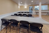 This single-floor multipurpose hall offers a clean, well-appointed space designed to accommodate a variety of gatherings, from meetings and classes to banquets and community events. The open floor plan is anchored by polished light hardwood flooring that extends throughout, lending warmth and a sense of spaciousness to the entire area.

Structural white columns are positioned symmetrically across the room, adding an architectural character while subtly defining zones within the open layout. The walls are finished in a soft warm beige tone and are dressed with crisp white wainscoting panels that run the full perimeter, providing an elegant, classic detail that elevates the overall aesthetic.

Recessed ceiling lighting ensures the space is evenly and brightly illuminated, while wall-mounted sconces contribute a softer ambient glow. The ceiling is smooth and unobtrusive, keeping the atmosphere clean and professional.

The hall is currently configured with rows of long folding tables paired with black folding chairs, demonstrating its capacity to comfortably seat a sizeable group in a classroom or banquet-style arrangement. A secondary room or utility area is visible at one end, suggesting additional functional support space. The overall ambiance is tidy, neutral, and highly adaptable, making this hall well-suited for a wide range of events and uses.