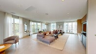 Located at 5919 N Kings Hwy in Alexandria, Virginia, this single-floor amenity space offers a generously sized, thoughtfully designed environment suited for lounging, socializing, and relaxation. The interior is defined by large floor-to-ceiling windows dressed with sheer white curtains that allow abundant natural light to filter throughout, creating a bright and airy atmosphere. Light-toned large-format tile flooring runs consistently across the space, lending a clean, cohesive aesthetic.

The main lounge area features an expansive sectional sofa arrangement anchored by a natural fiber area rug, complemented by additional seating clusters with rounded upholstered chairs and warm wood accent tables. Recessed lighting throughout the ceiling provides even, ambient illumination. A dedicated media area includes a built-in entertainment unit with light wood cabinetry and a wall-mounted display screen, with views to the outdoors just beyond.

Transitional spaces connect the interior to a well-appointed outdoor rooftop terrace, which features a covered pergola section with wood-slat ceiling detailing and an open-air seating arrangement. The terrace is furnished with wicker lounge chairs, striped sofas, and modern white tables, offering a comfortable outdoor retreat with open sky views. The overall ambiance throughout is contemporary, calm, and welcoming.