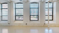 Located at 120 Valley St in Providence's West End, this single-floor loft unit reflects the building's industrial heritage through a thoughtfully converted interior. The main living area is an expansive, open space defined by tall ceilings and a row of large arched windows framed by painted white brick columns. These generational factory windows allow abundant natural light to pour in from multiple directions, offering views of surrounding greenery. Exposed metal ductwork runs overhead alongside track lighting, reinforcing the loft's industrial character.

The kitchen is efficiently laid out in an L-shape and features light maple cabinetry, granite countertops, a stainless sink, and a full suite of white appliances including a gas range, dishwasher, and refrigerator. The bathroom is clean and functional, finished with tile flooring, a full tub/shower combination, and a vanity with granite countertop detailing consistent with the kitchen. A utility area near the entry houses a water heater and additional storage behind louvered doors. Sliding doors within the main space provide flexible room division, enhancing the unit's adaptability for both living and working purposes.