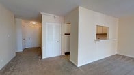 This single-floor residential space offers a straightforward and functional layout designed to maximize everyday comfort. The living area is finished with wall-to-wall carpeting in a neutral tone, contributing to a warm and quiet atmosphere throughout. A defining architectural feature is the partial partition wall that separates the living area from the kitchen, complete with a pass-through counter and upper cabinetry — an efficient design that maintains a sense of openness while clearly delineating the two spaces. The living area is further complemented by a set of classic louvered double closet doors, providing ample storage while adding a touch of traditional character to the room. A hallway leads further into the unit, suggesting additional rooms beyond the main living space. The textured ceiling and flush-mount lighting fixtures are consistent throughout, contributing to a cohesive and understated interior aesthetic. The overall layout is efficient and well-organized, with clearly defined zones for living, storage, and cooking. Neutral wall tones and light-colored trim work together to create a clean, bright environment that serves as a versatile backdrop for a variety of furnishing styles.