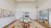 Located on Pine Forest Road in Pensacola, Florida, this single-story community amenity space offers a generously sized, open floor plan designed with a bright coastal aesthetic throughout. Soaring ceilings and abundant natural light set the tone from the moment you enter the main clubroom, where shiplap-clad walls, warm hardwood-style flooring, and large windows create an airy, welcoming atmosphere. A central lounge area features plush seating and a distinctive live-edge coffee table, flowing seamlessly into a spacious dining and gathering zone furnished with round tables and white Windsor-style chairs. The fully equipped kitchen serves as a social centerpiece, anchored by a large quartz-topped island with geometric patterned tile detailing and woven rattan bar seating. Woven globe pendant lights illuminate a striking deep-blue tile backsplash, white cabinetry, and stainless steel appliances. A dedicated fitness center offers an impressive array of strength and cardio equipment beneath high ceilings with ceiling fans and large windows overlooking the outdoors. Private glass-fronted office rooms provide quiet workspaces, while folding glass partition doors allow the various zones to connect or separate as needed. The cohesive coastal palette of whites, soft blues, and natural wood tones creates a polished yet relaxed community environment.