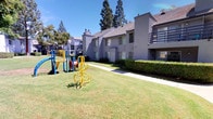 This residential community showcases a thoughtfully designed outdoor environment centered around expansive, well-kept communal lawns that serve as the heart of the complex. The buildings feature a classic stucco exterior in neutral gray tones, complemented by pitched rooflines and private balconies on the upper level, offering residents their own outdoor retreat with views of the surrounding greenery.

The grounds are a standout feature of the community, with wide stretches of manicured grass framed by mature trees that provide generous natural shade throughout the day. Winding concrete pathways meander through the landscape, connecting the various buildings and outdoor areas in a pedestrian-friendly layout. Neatly trimmed hedgerows line the building perimeters, adding a layer of greenery and privacy at ground level.

A dedicated children's playground is situated centrally within the communal lawn area, equipped with a colorful play structure including slides and climbing elements, as well as a spinning ride. Decorative mosaic seating elements are also present, adding an artistic touch to the outdoor communal space. Mature shade trees are strategically placed throughout, enhancing the park-like atmosphere. The overall ambiance is open, inviting, and community-oriented, making the outdoor spaces ideal for recreation and relaxation under bright, sunny skies.