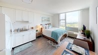 Located at 1221 Ala Kapuna Street in Honolulu, Hawaii, this well-appointed studio unit offers an efficient and thoughtfully designed open-plan layout on a single floor. Wood-look flooring runs throughout, providing warmth and visual continuity across all areas of the space.

The main living area seamlessly integrates sleeping, dining, and lounging functions. A full-sized bed serves as the focal point, positioned to take advantage of the expansive floor-to-ceiling windows that flood the room with natural light and frame sweeping panoramic views of the Honolulu cityscape and surrounding landscape. A compact dining table with upholstered chairs sits alongside a media console with a flat-screen TV, accented by coastal-inspired wall art that complements the unit's airy aesthetic.

The kitchenette is neatly integrated into the space, featuring white cabinetry, a full-sized refrigerator, electric range, microwave, and sink — providing all essential cooking amenities in a compact and functional arrangement.

The bathroom includes a walk-in shower with a curtained enclosure and a built-in shelf, finished in clean, neutral tones. Adjacent to the bathroom is a dedicated walk-in closet outfitted with wire shelving, offering generous storage for a studio unit.

Overall, the space presents a clean, modern, and coastal-inspired atmosphere, making efficient use of its layout while maximizing natural light and views.