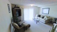 This two-floor residence offers a welcoming and functional living environment. The living room, pictured here, is a well-proportioned space finished with wall-to-wall carpeting that adds warmth underfoot. A large sliding glass door fitted with vertical blinds serves as the room's primary light source, flooding the interior with natural light and hinting at outdoor access. The textured ceiling adds a classic residential character to the space.

The living room is thoughtfully arranged to accommodate both entertainment and relaxation. A substantial dark wood entertainment armoire anchors one side of the room, housing a television and offering additional storage behind its cabinet doors. Decorative vessels displayed on top lend a personalized touch. On the opposing side, a light-toned sofa with accent pillows pairs with a glass-top coffee table supported by a wrought iron frame, creating an inviting seating arrangement. Wicker armchairs with cushioned seats and matching ottomans complete the furniture grouping, adding a casual, layered aesthetic. Framed wall art and lush indoor plants round out the space, contributing to a lived-in, comfortable ambiance throughout this multi-level home.