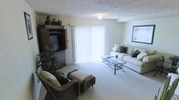 This two-floor residence offers a welcoming and functional living environment. The living room, pictured here, is a well-proportioned space finished with wall-to-wall carpeting that adds warmth underfoot. A large sliding glass door fitted with vertical blinds serves as the room's primary light source, flooding the interior with natural light and hinting at outdoor access. The textured ceiling adds a classic residential character to the space.

The living room is thoughtfully arranged to accommodate both entertainment and relaxation. A substantial dark wood entertainment armoire anchors one side of the room, housing a television and offering additional storage behind its cabinet doors. Decorative vessels displayed on top lend a personalized touch. On the opposing side, a light-toned sofa with accent pillows pairs with a glass-top coffee table supported by a wrought iron frame, creating an inviting seating arrangement. Wicker armchairs with cushioned seats and matching ottomans complete the furniture grouping, adding a casual, layered aesthetic. Framed wall art and lush indoor plants round out the space, contributing to a lived-in, comfortable ambiance throughout this multi-level home.