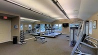 This fitness center is designed to support a comprehensive workout experience within a single, open-plan floor. The space features vaulted ceilings that contribute to an airy, expansive atmosphere, complemented by large windows along the walls that flood the room with natural light and provide pleasant outdoor views. The flooring is finished with durable, cushioned carpet suitable for fitness use.

Cardio training is well-represented with a row of treadmills positioned to face the windows, allowing users to enjoy the view during their workouts. A wall-mounted flat-screen television adds an entertainment option for those using the cardio equipment. Along one full wall, large mirrors visually expand the space and allow for proper form checks during exercise.

The free weights section includes a well-stocked dumbbell rack offering a range of weights, paired with an adjustable workout bench ideal for a variety of strength exercises. A multi-function cable and strength training machine is also present, rounding out the resistance training options.

Overhead, industrial-style lighting fixtures provide consistent, even illumination throughout the space. The warm, neutral-toned walls create a clean and focused environment. Overall, this fitness center is a functional and well-lit facility designed to accommodate a broad range of fitness needs.