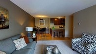 This single-floor residence showcases an open-concept layout that connects the living room, dining area, and kitchen in one cohesive space. The living area is anchored by a plush area rug and comfortable seating, accented with decorative throw pillows in warm tones of orange and neutral patterns. A sleek coffee table serves as a central focal point.

The dining area transitions naturally from the living space, featuring a bar-height table paired with stylish high-back chairs, illuminated by a modern pendant light overhead. A framed landscape artwork adorns the warm taupe-colored wall, adding visual interest.

The kitchen is appointed with rich dark-stained cabinetry, light-toned countertops, and stainless steel appliances, including a range and microwave. A breakfast bar with two bar stools provides additional seating and defines the kitchen's boundary. The warm hardwood-style flooring in the kitchen and dining area contrasts nicely with the carpeted living space, creating distinct yet connected zones. The overall ambiance is warm, modern, and functional.