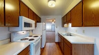 Located at 1559 Regent Ave in Springfield, Ohio, this single-floor unit offers a straightforward, functional layout spread across several interconnected rooms. The galley-style kitchen is well-equipped, featuring warm oak cabinetry on both sides, white appliances including a range, over-the-range microwave, and refrigerator, along with a double stainless steel sink set into light-toned countertops. Hardwood flooring runs throughout the kitchen, adding a classic touch. A secondary view of the kitchen reveals a back door with a window, providing additional natural light. The open living area is generously sized, with neutral carpet, a ceiling fan with light fixture, and baseboard heating. The layout flows naturally between the living space and adjacent rooms, as shown in the floor plan. Bedrooms are carpeted and bright, each featuring baseboard heating and windows with blinds that allow natural light. The bathroom is equipped with a full tub and shower surround, hardwood flooring, and a vanity with an oak cabinet base, light countertop, mirrored medicine cabinet, and lighted bar above. The overall aesthetic is clean and practical throughout.