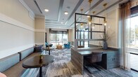 Alta River Oaks presents a thoughtfully designed single-floor amenity and leasing space that seamlessly blends contemporary style with functional versatility. The entrance greets visitors with a branded entryway featuring glass double doors, patterned tile flooring, and an elegant reception desk accented by gold pendant lighting and a mirrored open-frame divider, setting a polished first impression.

The open lounge area is anchored by plush teal velvet armchairs, a rustic wood block coffee table, and floor-to-ceiling windows that bathe the space in natural light while offering views of the tree-lined streetscape outside. A patterned, multi-tone carpet runs throughout, complementing the warm neutral palette of the walls and tray ceilings fitted with recessed and pendant lighting.

A communal meeting area features a solid wood dining table surrounded by mid-century modern chairs, exposed Edison-bulb pendants, and a sliding barn door that adds rustic charm. Adjacent to this is a dedicated conference room enclosed by glass-framed partitions, furnished with a natural wood conference table, rolling upholstered chairs, a wall-mounted display screen, and textured wallcovering that lends the room an intimate, professional atmosphere. A gold drum chandelier overhead adds a refined finishing touch throughout.