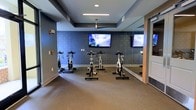 This single-floor spin studio is purpose-built for cycling workouts, offering a focused and well-equipped fitness environment. The room is finished with deep blue-gray textured walls that contribute to an energetic yet focused atmosphere. A tray ceiling with recessed lighting and a centrally mounted ceiling fan provides both adequate illumination and air circulation during intense workouts. The carpeted flooring offers a practical surface that anchors the multiple stationary cycling bikes arranged in a row for group or individual sessions. Two wall-mounted flat-screen televisions are positioned at the front of the room, allowing riders to follow workout content or entertainment during sessions. A large floor-to-ceiling mirror along one wall visually expands the space and allows users to monitor their form while cycling. The room is accessed through a standard door visible on the right side. Overall, the studio maintains a clean, modern aesthetic with a layout optimized for spin classes, combining functional fitness equipment with thoughtful design elements suited to an active, community-oriented fitness facility.