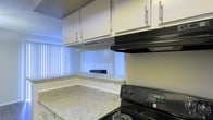 This single-floor space includes a well-appointed kitchen that serves as a central feature. The kitchen is outfitted with white upper cabinetry equipped with sleek bar-pull hardware, providing ample storage. Granite-style countertops in light tones with subtle veining add a polished look to the workspace. A black electric range with a matching overhead ventilation hood anchors the cooking area, offering a practical layout for meal preparation. A pass-through window connects the kitchen to an adjacent area, enhancing the sense of openness and allowing natural light to filter through. Vertical blinds along the window diffuse light while maintaining privacy. The neutral wall tones and textured ceiling complement the overall clean, functional aesthetic of the space. The kitchen's L-shaped counter configuration maximizes usable prep space, making it both practical and visually organized.