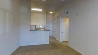 This single-story space features an open-concept design that visually connects the living area to the kitchen. The kitchen is defined by a raised peninsula with a dark countertop, providing a natural divide between the cooking and living spaces. White cabinetry lines the kitchen walls, complemented by built-in appliances including a microwave and refrigerator. Fluorescent overhead lighting illuminates the kitchen workspace, while a flush-mount ceiling fixture serves the adjacent areas. The living area is finished with wall-to-wall carpet in a neutral tone, contributing to a warm and comfortable atmosphere. Smooth, light-colored walls throughout give the space a clean and understated appearance. A hallway visible from the main living area leads to additional rooms, with white doors offering access to private spaces beyond. HVAC vents are integrated into the upper walls, ensuring climate control throughout. The overall layout is functional and straightforward, with a neutral color palette that provides a versatile backdrop for a variety of furnishing styles.
