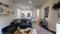 This contemporary single-floor residence showcases a thoughtfully designed open-concept layout that seamlessly connects the living, dining, and kitchen areas. The living room is anchored by a plush tufted sectional sofa in a rich charcoal tone, complemented by decorative throw pillows and a gold-accented coffee table that adds a warm, modern touch. Warm-toned wood-look flooring flows consistently throughout the space, enhancing the cohesive feel of the interior.

The kitchen is outfitted with stainless steel appliances, including a refrigerator, microwave, and range, all set against crisp white cabinetry with under-cabinet lighting. A peninsula with seating provides a casual dining option and reinforces the open flow between spaces. A compact dining nook with a round table and industrial-style chairs offers an additional spot for meals or work.

Recessed lighting throughout ensures the space is well-lit, while neutral gray walls provide a versatile backdrop for various décor styles. A hallway visible from the main living area leads to additional rooms and a bathroom, completing this well-appointed and efficiently organized residence. The overall ambiance is clean, modern, and welcoming.