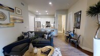 This contemporary single-floor residence showcases a thoughtfully designed open-concept layout that seamlessly connects the living, dining, and kitchen areas. The living room is anchored by a plush tufted sectional sofa in a rich charcoal tone, complemented by decorative throw pillows and a gold-accented coffee table that adds a warm, modern touch. Warm-toned wood-look flooring flows consistently throughout the space, enhancing the cohesive feel of the interior.

The kitchen is outfitted with stainless steel appliances, including a refrigerator, microwave, and range, all set against crisp white cabinetry with under-cabinet lighting. A peninsula with seating provides a casual dining option and reinforces the open flow between spaces. A compact dining nook with a round table and industrial-style chairs offers an additional spot for meals or work.

Recessed lighting throughout ensures the space is well-lit, while neutral gray walls provide a versatile backdrop for various décor styles. A hallway visible from the main living area leads to additional rooms and a bathroom, completing this well-appointed and efficiently organized residence. The overall ambiance is clean, modern, and welcoming.