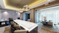 This refined single-floor professional office space blends high-end design with functional workspaces, creating an environment that is both impressive and purposeful. The centerpiece of the space is a dramatically backlit marble conference table, its white and grey veining illuminated from within, surrounded by plush leather chairs. Above, a series of geometric crystal-glass pendant lights hang from a coffered ceiling accented with warm ambient uplighting, adding a layer of luxury to the workspace. Flanking the main area are built-in dark wood bookshelves, thoughtfully curated with books and decorative objects, lending a scholarly sophistication to the environment. A dedicated library nook features floor-to-ceiling shelving on three walls, paired with two upholstered accent chairs and a round side table — ideal for focused reading or quiet meetings. Glass-walled private offices line the perimeter, offering visual connectivity while maintaining acoustic privacy. Sheer white curtains diffuse natural light throughout these offices, contributing to a bright and airy feel. Decorative crown molding and warm wood column detailing frame transitions between spaces, reinforcing the space's polished, professional character. The overall ambiance is one of understated elegance, designed to impress clients and inspire productivity.