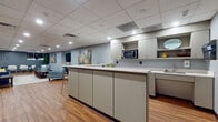 Located in Hartford, CT, this thoughtfully designed single-floor space functions as a versatile common area, blending comfort with a clean, contemporary aesthetic. The layout flows seamlessly from a fully equipped kitchenette to an open lounge area, making it well-suited for both casual socializing and organized gatherings.

The kitchen area is outfitted with an expansive run of light-toned cabinetry — both upper and lower — featuring ample storage and generous countertop space with a light quartz-style surface. A built-in microwave, sink, and a water dispenser contribute to the room's practical functionality. Warm hardwood-style flooring extends throughout the entire space, tying the zones together with a cohesive, inviting look.

The lounge area is furnished with a mix of seating options, including a navy blue sofa accented with yellow throw pillows, a chartreuse accent chair, and a striped armchair, all arranged around a central area rug. A wall-mounted flat-screen TV is positioned above a sleek linear fireplace, creating a natural focal point. Abstract artwork displayed on the blue-gray walls adds a layer of character and visual warmth.

The space is illuminated by recessed lighting set within a drop tile ceiling, providing consistent, even lighting throughout. A glass-partitioned room is visible along one wall, offering a defined private area within the larger open-plan layout. Overall, the space presents a welcoming, well-maintained environment designed for comfort and community use.