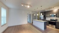 This single-floor, 3-bedroom, 2-bathroom residence offers a thoughtfully designed open-concept layout that connects the living area and kitchen in one cohesive space. The living area features light-toned hardwood-style flooring and crisp white walls, creating a bright and airy atmosphere. Track lighting runs along the ceiling, providing flexible illumination throughout the main living space.

The kitchen is a standout feature, anchored by a sleek island with a light stone countertop and an undermount sink. Pendant lighting hangs above the island, adding both function and style. Dark cabinetry contrasts elegantly with the lighter countertops and walls, while stainless steel appliances, including a French door refrigerator, complete the modern aesthetic. Additional cabinetry lines the perimeter of the kitchen, offering generous storage.

A doorway visible from the main living area leads to the private quarters of the residence, maintaining a clear separation between shared and personal spaces. The overall design emphasizes clean lines, neutral tones, and contemporary finishes throughout, making it a versatile and functional living space.
