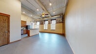 Located at 101 N Kansas Ave in downtown Topeka, this 2-bedroom, 2-bathroom loft-style residence blends historic industrial character with modern finishes. The open-concept living and kitchen area is immediately striking, featuring soaring ceilings with exposed wooden beams, visible ductwork, and a ceiling fan that emphasizes the generous vertical space. Exposed brick accent walls line the upper perimeter, complementing large windows that invite abundant natural light. The kitchen is equipped with warm wood cabinetry, a tiled backsplash, granite countertops, stainless steel appliances, pendant lighting, and a central island with bar seating. Both bedrooms offer plush patterned carpet, tall ceilings, and natural light from large windows framed by the building's original brick. The spacious primary bedroom provides ample room for comfortable furnishings. Each bathroom is appointed with wood-framed vanities, granite countertops, and walk-in showers with tiled surrounds. A dedicated closet and two hallways provide practical storage and circulation throughout the unit. The overall aesthetic is a refined industrial loft experience, marrying the character of a historic building with thoughtfully updated contemporary interiors.