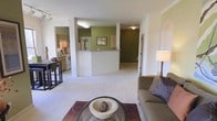 This single-floor residence offers an open, flowing layout that connects the living area, kitchen, and hallway in a cohesive arrangement. The living room serves as the central gathering space, featuring light neutral carpeting, a glass-topped coffee table, and a comfortable upholstered sofa accented with decorative pillows. A floor lamp provides warm ambient lighting, complementing the overall inviting atmosphere.

A half-wall partition separates the kitchen from the main living area, creating a semi-open pass-through design that maintains visual connectivity while defining each zone. The kitchen is equipped with wooden cabinetry, a stainless steel range, and standard appliances, offering a functional and practical cooking space. The green accent walls in both the kitchen and hallway add a distinct pop of color that contrasts with the white walls and trim throughout.

A hallway extending from the main living space leads toward additional rooms, lined with the same neutral carpeting that runs throughout the residence. The arched doorway detail visible on one wall adds a subtle architectural element. Overall, the space presents a clean, functional layout with warm color accents and consistent flooring that ties each area together seamlessly.