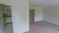 This single-floor space presents a straightforward and functional layout. The main room is finished with wall-to-wall carpeting in a neutral beige tone, complemented by smooth, white-painted walls that lend a clean and bright atmosphere throughout. A textured ceiling with a central ceiling vent contributes to the room's modest, practical character. A partial interior wall creates a subtle division within the space, adding a degree of architectural definition without fully enclosing the area. An open doorway leads to an adjoining room featuring light-colored tile flooring, suggesting a utility, bathroom, or transitional space. The overall color palette remains consistently neutral, with white walls and light flooring tones creating a cohesive, understated aesthetic. Electrical outlets and a light switch are visible along the walls, indicating the space is move-in ready. The layout is open and unobstructed, making it adaptable for a variety of uses. Overall, the space offers a clean, blank-canvas environment suited for straightforward occupancy or customization.