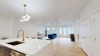 This thoughtfully designed single-floor residence offers a seamless blend of modern aesthetics and functional living across a generous footprint. The open-concept kitchen and living area serves as the heart of the home, anchored by a striking oversized waterfall-edge island topped in white marble-look quartz and lined with navy velvet barstools featuring gold accents. The kitchen is equipped with two-tone cabinetry — crisp white upper cabinets paired with deep navy lowers — stainless steel appliances including a French door refrigerator, a professional-grade gas range with a chimney hood, and a bold globe chandelier overhead. Light hardwood flooring flows throughout, enhancing the bright, airy atmosphere created by white walls and abundant natural light from multiple windows. The living area features a ceiling fan, direct access to the outdoors via French doors, and generous room to entertain. The spacious primary bedroom accommodates a large bed with ease and benefits from dual windows and its own ceiling fan. It connects directly to a well-appointed en-suite bathroom with navy shaker-style double vanity, gold hardware, an illuminated mirror, and large-format tile flooring. A dedicated walk-in closet with wire shelving provides ample storage. A second bathroom and a dedicated laundry room round out this well-equipped residence.