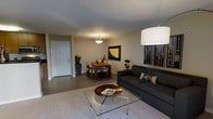 This single-floor residence offers a thoughtfully designed open-concept layout that seamlessly blends the living and dining areas into a cohesive, inviting space. The living area is anchored by a sleek charcoal sofa adorned with accent pillows, complemented by a glass-topped coffee table that adds a touch of modern elegance. A large drum pendant light illuminates the space with a warm glow, enhancing the room's comfortable ambiance. The walls are decorated with framed artwork, including a striking cityscape print and a bold yellow abstract piece, lending a curated, contemporary feel throughout. The dining area features a warm wood table surrounded by upholstered chairs, centered beneath a pendant light fixture. Potted greenery is thoughtfully placed throughout, adding natural life and color to the neutral palette of soft grays and warm whites. The flooring transitions smoothly between tile and carpet, subtly defining each functional zone. A solid white interior door near the entryway provides access to additional areas of the home. Overall, the space conveys a clean, modern sensibility with warm, welcoming touches that make it both functional and aesthetically refined.