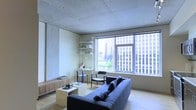This contemporary single-floor space showcases a refined minimalist aesthetic defined by an exposed concrete ceiling that lends an industrial yet sophisticated character to the interior. The living area is thoughtfully arranged, featuring a streamlined sofa, a low-profile wooden coffee table, and a compact wall-mounted shelving unit that doubles as a workspace. Light wood flooring adds warmth and contrast to the cool concrete overhead, creating a balanced and inviting atmosphere throughout.

Large picture windows serve as a defining architectural feature, flooding the space with natural light while framing impressive views of the surrounding urban skyline. The cityscape visible through the glass — with its mix of high-rise buildings and greenery — becomes a dynamic backdrop that enhances the overall ambiance of the interior.

The layout is open and efficient, with furnishings carefully selected to complement the clean lines of the architecture. A dedicated desk area with open shelving provides a functional workspace integrated seamlessly into the living environment. The overall design philosophy emphasizes simplicity, natural materials, and a strong connection to the vibrant city surroundings, making this a compelling urban space suited for both focused work and relaxed living.
