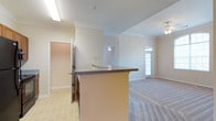 This single-floor residence showcases an open-concept design that seamlessly connects the kitchen and living areas. The kitchen features a peninsula with a dark laminate countertop, providing both additional workspace and a natural dividing point between the cooking and living zones. The flooring transitions from tile in the kitchen to plush carpet in the living area, offering a clear distinction between functional spaces. Crown molding runs throughout the home, adding a refined architectural detail to the neutral, light-colored walls. The living area benefits from natural light streaming through a glass-paneled exterior door, complemented by a ceiling fan overhead. A secondary hallway leads to additional rooms, hinting at a practical and well-organized floor plan. The textured ceiling and recessed lighting in the kitchen add subtle depth to the space. Overall, the residence presents a clean, neutral palette with warm undertones, creating a welcoming and functional environment suited to everyday living.