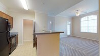 This single-floor residence showcases an open-concept design that seamlessly connects the kitchen and living areas. The kitchen features a peninsula with a dark laminate countertop, providing both additional workspace and a natural dividing point between the cooking and living zones. The flooring transitions from tile in the kitchen to plush carpet in the living area, offering a clear distinction between functional spaces. Crown molding runs throughout the home, adding a refined architectural detail to the neutral, light-colored walls. The living area benefits from natural light streaming through a glass-paneled exterior door, complemented by a ceiling fan overhead. A secondary hallway leads to additional rooms, hinting at a practical and well-organized floor plan. The textured ceiling and recessed lighting in the kitchen add subtle depth to the space. Overall, the residence presents a clean, neutral palette with warm undertones, creating a welcoming and functional environment suited to everyday living.