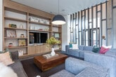 Located in the vibrant Halcyon development in Alpharetta, Georgia, this thoughtfully designed single-floor space blends modern aesthetics with functional elegance. The open-concept layout showcases soaring ceilings that amplify the sense of volume and airiness throughout.

The kitchen area is a standout feature, outfitted with sleek light wood cabinetry, expansive quartz countertops, and stainless steel appliances including a wall oven, dishwasher, and undercounter refrigerator. Illuminated floating shelves line the walls, providing both display and storage while adding a warm, curated ambiance. A deep undermount sink anchors the continuous countertop run, lending a clean, professional aesthetic.

The lounge area offers a welcoming, relaxed atmosphere centered around a generous modular sectional sofa positioned on large-format tile flooring that transitions seamlessly to light hardwood. The soaring walls are adorned with bold black-and-white photographic artwork, adding a cosmopolitan character to the space. A floor-to-ceiling built-in shelving unit in natural wood tones provides generous storage and display space, complemented by a pendant light fixture overhead. The overall palette of soft whites, warm woods, and cool grays creates a cohesive, contemporary environment ideal for gathering and relaxation.