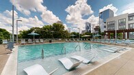 Located at 1015 Nelson Merry Street in Nashville's vibrant Gulch neighborhood, this outdoor amenity space delivers a resort-caliber experience in the heart of the city. At its center sits a generously sized swimming pool with clear turquoise water, framed by an extensive sun deck lined with lounge chairs and umbrellas for ample relaxation space. Overhead, striking wood-beam pergolas provide partial shade throughout the deck area, adding a warm architectural contrast to the surrounding contemporary multi-story building. A raised terrace runs along the perimeter, enclosed by sleek glass-panel railings that preserve unobstructed sightlines. From this elevated walkway, residents enjoy sweeping views of Nashville's downtown skyline, with glass high-rises rising above a lush canopy of mature trees — a compelling blend of urban energy and natural greenery. Integrated landscaping, including ornamental grasses and plantings, adds a polished, resort-like finish to the space. The layout is thoughtfully designed to offer distinct zones for active swimming, lounging in the sun, and relaxing in shaded seating areas, making it a versatile outdoor destination year-round. The overall ambiance balances modern design with an inviting, leisurely atmosphere befitting its prime Nashville location.