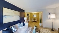 This single-floor residence features two bedrooms and one bathroom, offering a thoughtfully designed layout that maximizes both functionality and style. The open-concept living and dining area serves as the heart of the home, creating a seamless flow between spaces. The living room is anchored by a bold navy accent wall, which provides a striking contrast against the crisp white ceiling and warm wood-look flooring that extends throughout the unit. A large piece of abstract wall art adds a sophisticated touch to the space.

The dining area is defined by warm yellow walls and illuminated by a modern multi-bulb chandelier, creating an inviting atmosphere for gatherings. A glass-top dining table with upholstered chairs completes the setting. Just beyond, the kitchen is partially visible, featuring bar-height seating that offers a casual dining alternative.

The light-toned flooring runs consistently throughout, unifying the living, dining, and kitchen areas. The overall design palette — combining navy, gold, and neutral tones — creates a cohesive, contemporary aesthetic. The layout makes efficient use of the generous square footage, providing ample room for comfortable everyday living.