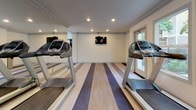 Located at 240 Burt St in Santa Rosa, California, this fitness center offers a well-appointed, single-floor exercise facility designed to support a complete workout routine. The space features a distinctive striped hardwood-style floor in alternating light and dark tones, adding a dynamic visual element throughout the room. Recessed lighting illuminates the interior evenly, complemented by ample natural light streaming through multiple windows and a glass-paneled exterior door. Full-length mirrored walls on one side expand the sense of space and allow users to monitor their form during workouts. The cardio section is equipped with multiple treadmills and an elliptical machine, while the strength training area includes an adjustable workout bench and a vertical knee raise and dip station. A wall-mounted flat-screen television provides entertainment during exercise sessions. Crown molding details along the ceiling add a refined architectural touch to the otherwise clean, neutral-toned walls. The exterior-facing windows offer views of a nearby swimming pool and lush surrounding greenery, creating an inviting and motivating atmosphere. A waste bin and posted facility guidelines near the entrance reflect thoughtful, practical amenities. The layout is open and uncluttered, providing users with generous floor space for stretching or additional exercises.
