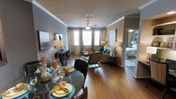 This single-floor residence showcases an open-concept layout that seamlessly connects the dining and living areas, creating a cohesive and functional space. Rich hardwood flooring runs throughout, lending warmth and a sense of continuity between rooms. The dining area is positioned at the front of the space, accommodating a circular glass-top table surrounded by dark upholstered chairs, ideal for entertaining. Crown molding adds an elegant architectural detail along the ceiling line, while a ceiling fan with a light fixture provides both comfort and ambient illumination to the living area. The living room benefits from multiple windows dressed with floor-length draping curtains, allowing natural light to fill the space. Neutral and grey-toned walls provide a versatile backdrop for furnishings. A media console anchors one wall of the living area, complemented by coordinated seating arrangements. A doorway visible from the main living space leads to a bedroom, hinting at a well-organized floor plan that balances private and shared living zones. Overall, the space presents a polished, move-in-ready aesthetic with thoughtful design details throughout.