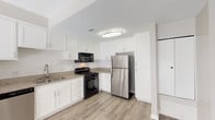 Located at 22900 Oak Ridge Dr in Santa Clarita, CA, this single-story unit offers a clean, modern aesthetic throughout. The kitchen is a standout feature, showcasing crisp white cabinetry, granite-style countertops, a subway tile backsplash, and stainless steel appliances including a refrigerator and dishwasher, complemented by a black gas range and over-the-range microwave. Light wood-style vinyl plank flooring runs throughout the main living areas, lending warmth and a cohesive look to the space. The open layout connects the kitchen to a generously sized living area, which is bathed in natural light from a large sliding glass door fitted with vertical blinds, offering access to an outdoor area. The bedroom features plush neutral carpeting and a window with blinds that allows for soft, diffused light. The bathroom is well-appointed with white cabinetry, granite-style countertops, a vessel sink, and a tub-shower combination with white tile surround and a safety grab bar. Brushed nickel hardware accents are found throughout the bathroom, adding a polished finishing touch. The overall color palette of warm grays and whites creates a bright, clean, and inviting atmosphere throughout the entire unit.