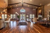 This single-story community clubhouse and leasing center is defined by its dramatic architectural presence and thoughtfully designed interior spaces. The grand entrance foyer welcomes visitors with soaring vaulted ceilings accented by dark wood beam detailing, an arched transom window flooding the space with natural light, and rich, warm hardwood flooring that flows continuously throughout. A tall, sculptural indoor plant anchors the entryway, setting an inviting tone.

The expansive main hall opens into multiple distinct zones. A stylish lounge area features plush seating, decorative spherical wall art, and large windows with views to the outdoors. A sophisticated fireplace lounge anchors one end of the space, showcasing a floor-to-ceiling tiled fireplace surround with built-in bench seating, curated artwork, and a bold purple area rug beneath a contemporary round coffee table.

The leasing area includes a sleek reception counter with recessed ceiling detail, mirrored wall panels, and an intimate seating nook for consultations. Adjacent to the main space is a fully appointed display kitchen featuring dark espresso cabinetry, granite countertops with a curved island, white subway tile backsplash, and stainless steel appliances. Throughout, the space balances contemporary design with warm, inviting finishes.