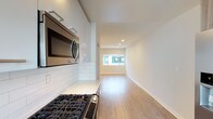 This single-floor residence offers a clean, modern aesthetic defined by an open-concept layout that seamlessly connects the kitchen and living area. The kitchen is thoughtfully appointed with sleek, high-gloss white cabinetry, classic white subway tile backsplash, and stainless steel appliances including a gas range and an over-the-range microwave. Light-toned countertops complement the bright, airy palette of the space. The flooring throughout is a warm light hardwood, lending a sense of continuity and warmth as it flows from the kitchen into the living area. Recessed lighting in the ceiling provides even, ambient illumination, while a wide window at the far end of the living space invites natural light deep into the interior. The walls are finished in a crisp, neutral white, enhancing the open and uncluttered feel of the space. The overall ambiance is fresh, contemporary, and move-in ready, offering a versatile canvas suited to a variety of interior styles. The open layout maximizes the sense of space, making the residence feel bright and inviting throughout.
