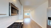 This single-floor residence offers a clean, modern aesthetic defined by an open-concept layout that seamlessly connects the kitchen and living area. The kitchen is thoughtfully appointed with sleek, high-gloss white cabinetry, classic white subway tile backsplash, and stainless steel appliances including a gas range and an over-the-range microwave. Light-toned countertops complement the bright, airy palette of the space. The flooring throughout is a warm light hardwood, lending a sense of continuity and warmth as it flows from the kitchen into the living area. Recessed lighting in the ceiling provides even, ambient illumination, while a wide window at the far end of the living space invites natural light deep into the interior. The walls are finished in a crisp, neutral white, enhancing the open and uncluttered feel of the space. The overall ambiance is fresh, contemporary, and move-in ready, offering a versatile canvas suited to a variety of interior styles. The open layout maximizes the sense of space, making the residence feel bright and inviting throughout.