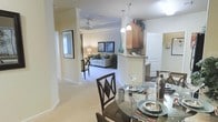 This single-story residence offers an open-concept floor plan that seamlessly integrates the dining, kitchen, and living spaces into one cohesive environment. The dining area, positioned in the foreground, features a round glass-top table with elegant dark wood chairs, creating a refined yet approachable setting for meals. A peninsula-style kitchen counter serves as a natural divider between the dining and kitchen areas, topped with light-colored surfaces and flanked by warm wood cabinetry above. Pendant lighting drops from the ceiling over the counter, adding focused illumination and a decorative touch.

The living area beyond is carpeted and comfortably appointed, anchored by a light gray sofa with accent pillows and framed artwork on the walls. A ceiling fan with a light fixture overhead provides both airflow and ambient light to the space. Decorative columns and archways add architectural interest and definition between zones without closing off the open layout.

Throughout the home, a soft, neutral color palette of cream and beige tones creates a warm, welcoming ambiance. Crown molding along the ceilings adds a refined finish, while natural light filtering in from surrounding windows keeps the interior bright and airy.