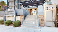 This single-floor common area serves as the primary entry and mailroom for the building. The exterior entrance makes a distinctive first impression with its natural stacked-stone surround framing a set of glass double doors. A covered overhead canopy provides shelter at the entry, while a concrete staircase with metal railings leads up to the doors, giving the approach a structured, defined character. A security intercom panel is mounted near the entry for controlled access.

Inside, the lobby is finished with neutral beige tiling and a drop ceiling fitted with recessed fluorescent lighting that keeps the space well-illuminated. A full wall of individual tenant mailboxes lines one side of the corridor, offering organized mail distribution for building residents. Along the opposite wall, a narrow wooden shelf desk accompanied by a blue upholstered chair provides a convenient spot for sorting mail or briefly reviewing materials. A small recycling or waste receptacle is positioned nearby. Interior doors provide access to additional areas of the building. The overall ambiance is clean, functional, and practical, designed to efficiently serve the day-to-day needs of building residents.