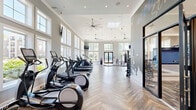 Located in Matthews, NC, this fitness facility offers an expansive, thoughtfully designed workout environment spread across a single floor. The space is defined by its dramatic soaring ceilings, which are accentuated by large, multi-pane windows arranged in stacked tiers along the walls, flooding the interior with abundant natural light and offering pleasant outdoor views of the surrounding greenery.

The herringbone-patterned wood-look flooring adds a refined, upscale aesthetic throughout, while oversized ceiling fans with sleek, modern profiles provide airflow and comfort during workouts. Pendant lighting and recessed fixtures further enhance the bright, airy ambiance.

The two exercise rooms are comprehensively equipped to support a wide range of training styles. Cardio enthusiasts will find a dedicated row of treadmills and elliptical machines, each outfitted with personal screens and positioned to face the large windows. Strength and functional training areas feature a multi-station cable machine, an Octagon functional training rig, a heavy punching bag, battle ropes, a flip tire, dumbbell racks with varied weights, and medicine balls. Wall-mounted televisions are positioned throughout for entertainment. A set of double glass doors opens directly to the outdoors, seamlessly connecting the interior training space to the exterior environment.