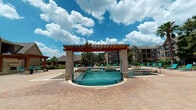 The Enclave in College Station, Texas offers a thoughtfully designed outdoor amenity space that serves as a central gathering hub for its community. The centerpiece is a resort-style swimming pool with a striking curved design, complemented by a separate hot tub and decorative water features. A bold wooden pergola with stone-clad columns stands over the pool area, adding architectural character while providing partial shade to the surrounding paver deck. Vibrant teal lounge chairs line the pool's edge, offering ample space for relaxation under the Texas sky. Adjacent to the pool, a covered outdoor seating area features a wood-beamed pergola strung with Edison-style lights, sheltering multiple patio tables and chairs — ideal for casual dining or socializing. A standout feature of the space is a substantial stacked-stone outdoor fireplace, adding a warm and inviting focal point to the patio. Lush landscaping, including tall palm trees and colorful flowering plants, frames the space and adds a tropical ambiance. The surrounding multi-story brick residential buildings provide a sense of community scale, while wrought iron fencing and gated access points ensure a secure and well-maintained environment throughout.