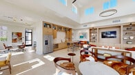 Located at 29986 Berea Road in Menifee, California, this single-story community amenity space is thoughtfully designed to serve as a versatile gathering and work hub. The interior is finished with large-format light tile flooring throughout, crisp white walls, and soaring ceilings that create a bright, airy atmosphere. Expansive clerestory windows and floor-to-ceiling black-framed sliding glass doors flood the space with natural light while offering direct views and access to an outdoor pool and patio area.

The main open-plan area seamlessly integrates multiple functional zones. A fully equipped kitchenette features warm wood cabinetry, a tile backsplash, stainless steel appliances including a full-size refrigerator, and a bar-height communal table with seating. Adjacent to the kitchen, a cozy lounge area offers built-in shelving, a mounted television, banquette seating with colorful accent pillows, and an assortment of comfortable chairs and sofas for relaxed socializing.

A dedicated conference and workspace room provides ample natural light through large windows on multiple walls, accommodating a long conference table, rolling office chairs, and casual seating arrangements — ideal for meetings or focused work sessions. The overall design palette of warm wood tones, neutral furnishings, and curated artwork creates a welcoming, contemporary ambiance throughout.