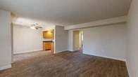 This single-floor unit offers a functional and straightforward layout designed to maximize usable living space. The expansive main living area is finished with wall-to-wall carpet in a warm tan tone and features a ceiling fan overhead. A structural beam subtly defines the space, while neutral gray walls provide a clean, versatile backdrop. The living area connects openly to the kitchen via a half-wall peninsula, creating a cohesive flow between the two spaces.

The galley-style kitchen is efficiently organized with warm honey-toned wood cabinetry lining both walls, dark laminate countertops, and recessed lighting above. White appliances, including a gas range, microwave, refrigerator, and dishwasher, are included. Tile flooring in a stone-like pattern completes the kitchen.

The unit includes multiple bedrooms, all carpeted and finished with neutral walls and white baseboards. One bedroom features a mirrored sliding closet and a window with natural light. Additional bedrooms offer similar sizing and understated finishes. The bathroom includes wood-look plank tile flooring, a dark-toned vanity, and a full tub-shower combination with white tile surround.