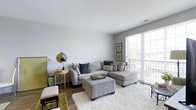 Located at 3443 Park Street in Grove City, Ohio, this single-floor residence combines modern design with comfortable, functional living. The living area serves as the centerpiece of the home, featuring neutral gray walls that create a clean, airy atmosphere. Large sliding glass doors flood the space with abundant natural light while providing direct access to a private outdoor balcony, offering pleasant views of the surrounding greenery. The room accommodates a generous sectional sofa arrangement anchored by a plush, textured area rug, giving the space a warm and inviting feel. Hardwood-style flooring runs throughout, adding warmth and a sense of continuity to the interior. The high, smooth ceilings enhance the open, uncluttered character of the layout, contributing to an overall sense of spaciousness. Thoughtful details such as built-in ventilation and carefully placed accent lighting reflect an attention to practicality and comfort. The design aesthetic throughout is contemporary yet approachable, blending soft tones with natural textures to create a welcoming environment. This Grove City residence offers a well-balanced living experience in a conveniently located neighborhood.