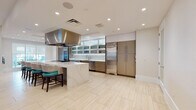 Located at 1231 Arya Way in Ocoee, Florida, this single-floor space is designed with a contemporary aesthetic and a strong emphasis on culinary functionality. The centerpiece is a spacious, professionally appointed kitchen finished in a neutral palette of white and warm taupe cabinetry. Upper cabinets feature frosted glass panel inserts, while under-cabinet lighting illuminates the striking marble-look quartz countertops and backsplash throughout. A large central island with a waterfall-edge quartz surface provides generous prep space and seating for multiple guests, outfitted with stylish teal and textured upholstered barstools. Premium stainless steel appliances include a high-capacity commercial-style range, an oversized refrigerator, a built-in wine cooler, and a dishwasher, all complemented by a bold stainless steel range hood overhead. Light-toned large-format tile flooring runs throughout, enhancing the bright, open atmosphere created by recessed ceiling lighting. A dedicated dining area adjacent to the kitchen features a wood-top table and cushioned seating, positioned alongside expansive floor-to-ceiling windows fitted with roller shades. These windows open to views of an outdoor patio furnished with seating and colorful landscaping, seamlessly connecting the interior to the exterior. Glass-panel interior doors with polished chrome hardware complete the refined, modern character of this well-appointed space.